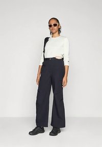 Balti marškiniai ilgomis rankovėmis, juodos juosmens kelnės su kišenėmis, juodi sportbačiai ir dideli akiniai nuo saulės.