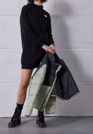 Persona con vestido tipo sudadera negro y botas negras, sosteniendo una chaqueta acolchada verde y negra contra un fondo de pared gris.