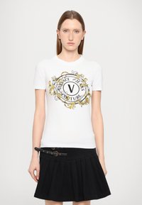 Valge puuvillane T-särk, mille esiküljel on must ja kuldne graafika "Versace Jeans Couture" ning dekoratiivsed barokkielemendid. Must volangseelik.