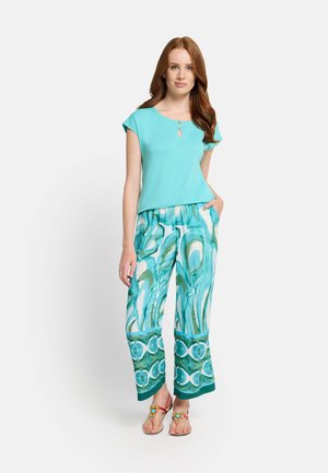 Woman with long red hair wearing a turquoise top and patterned wide-leg pants with sandals, standing with one hand in pocket.