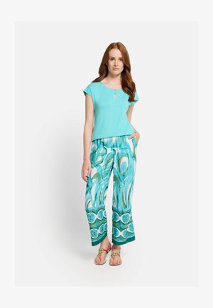 Woman with long red hair wearing a turquoise top and patterned wide-leg pants with sandals, standing with one hand in pocket.