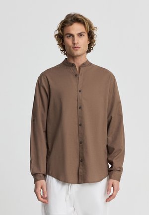 Giovane uomo con capelli ricci che indossa una camicia marrone con bottoni e colletto alla coreana e pantaloni bianchi, in piedi su uno sfondo semplice.