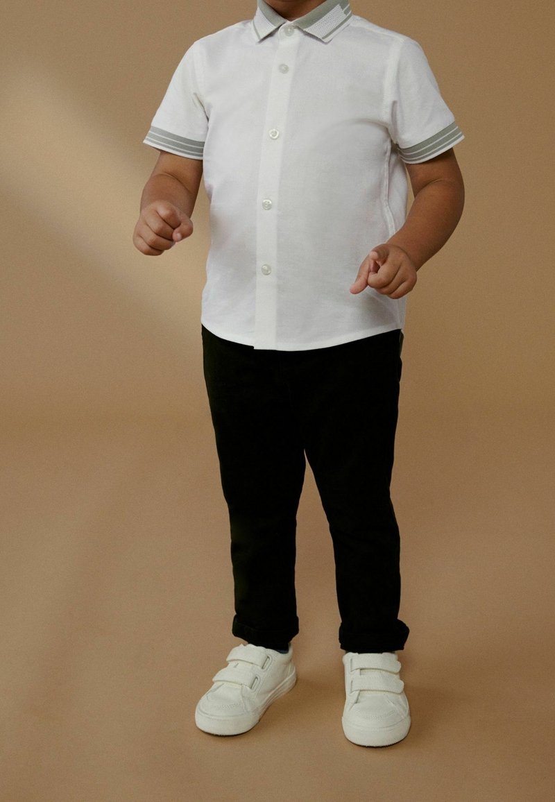 Enfant portant une chemise blanche à manches courtes boutonnée, un pantalon noir et des baskets blanches à scratch, debout devant un fond beige.