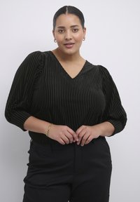 Černá, texturovaná blůza s vertikálními pruhy, výstřihem do V a balónovými rukávy. V kombinaci s černými kalhotami a zlatým náramkem.