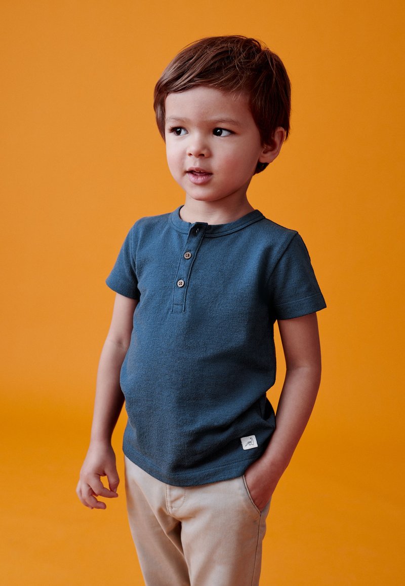 Jeune garçon aux cheveux bruns courts portant une chemise bleue boutonnée et un pantalon beige, debout devant un fond orange.