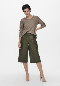 Brauner Strickpullover mit V-Ausschnitt, kombiniert mit olivgrünen weit geschnittenen Hosen und beigen High Heels. Das Model steht selbstbewusst mit einer Hand auf der Hüfte.