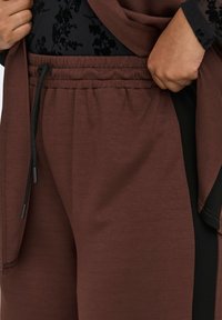 Brown jogger pants with an elastic waistband and black accents. The fabric appears soft with a slightly textured finish, featuring black drawstrings.