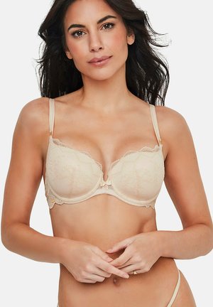 Vrouw met lang donker haar, gekleed in een beige kanten beha, handen zachtjes in elkaar gevouwen ter hoogte van de taille, neutrale achtergrond.