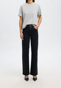 Graues Baumwoll-T-Shirt mit kurzen Ärmeln, kombiniert mit hochtaillierten, schwarzen weit geschnittenen Jeans und schwarzen spitzen Absatzschuhen, stehend vor einem neutralen Hintergrund.