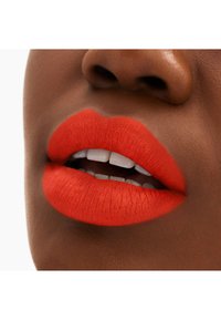 Rouge à lèvres mat rouge sur des lèvres pleines, contrastant avec un teint de peau foncée ; lignes fines visibles et texture lisse avec des dents blanches révélées.
