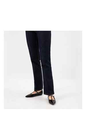 Personne portant un jean droit bleu foncé et des ballerines à bride en T noires à bout pointu sur fond blanc.