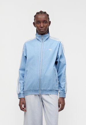 Femme portant une veste de survêtement bleu clair zippée avec des rayures blanches sur les manches et un pantalon de couleur claire, debout devant un fond uni.