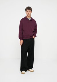 Burgundy polo shirt with a collar, black wide-leg trousers, and white sneakers with a tan sole. Casual fit, with long sleeves and relaxed design.