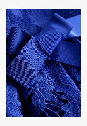 Material de dantelă texturată albastră cu o fundă mare din satin cu nervuri, legată în centru, evidențiind modele florale complicate și detalii fine de cusături.