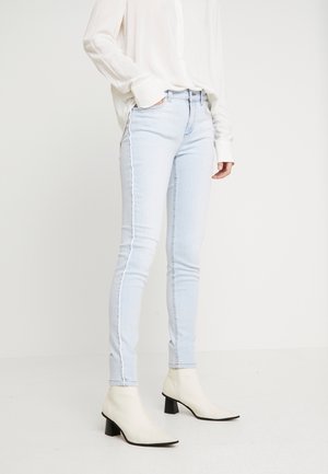 Femme portant un jean skinny bleu clair avec des coutures latérales effilochées, un chemisier blanc et des bottines blanches à talons blocs bas.