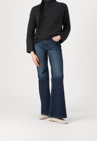 Personne portant un pull à col roulé gris foncé, un jean évasé bleu et des chaussures plates beiges, debout devant un fond blanc uni.