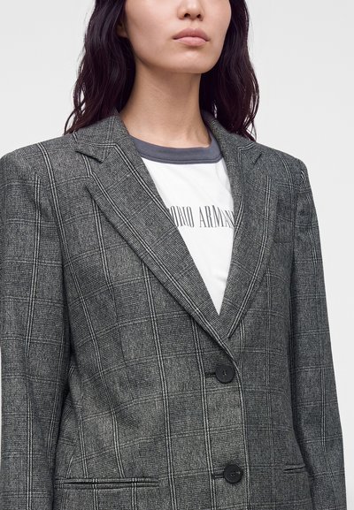 Personne portant un blazer à carreaux gris par-dessus un t-shirt blanc avec le logo Giorgio Armani partiellement visible sur un fond clair uni.