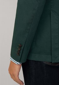 Blazer vert à finition texturée, présentant quatre boutons foncés sur le poignet et un col de chemise rayé visible en dessous.