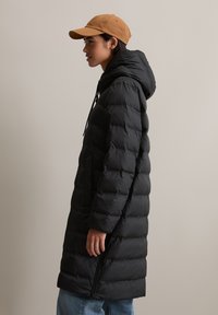 Svart pufferjakke med horisontalt quiltet mønster, hette og snor; kombinert med en beige caps og blå jeans; sideprofil.