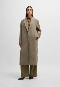Long beige coat with textured fabric, front pockets, and notched lapels, worn over a brown shirt and olive green wide-leg pants.