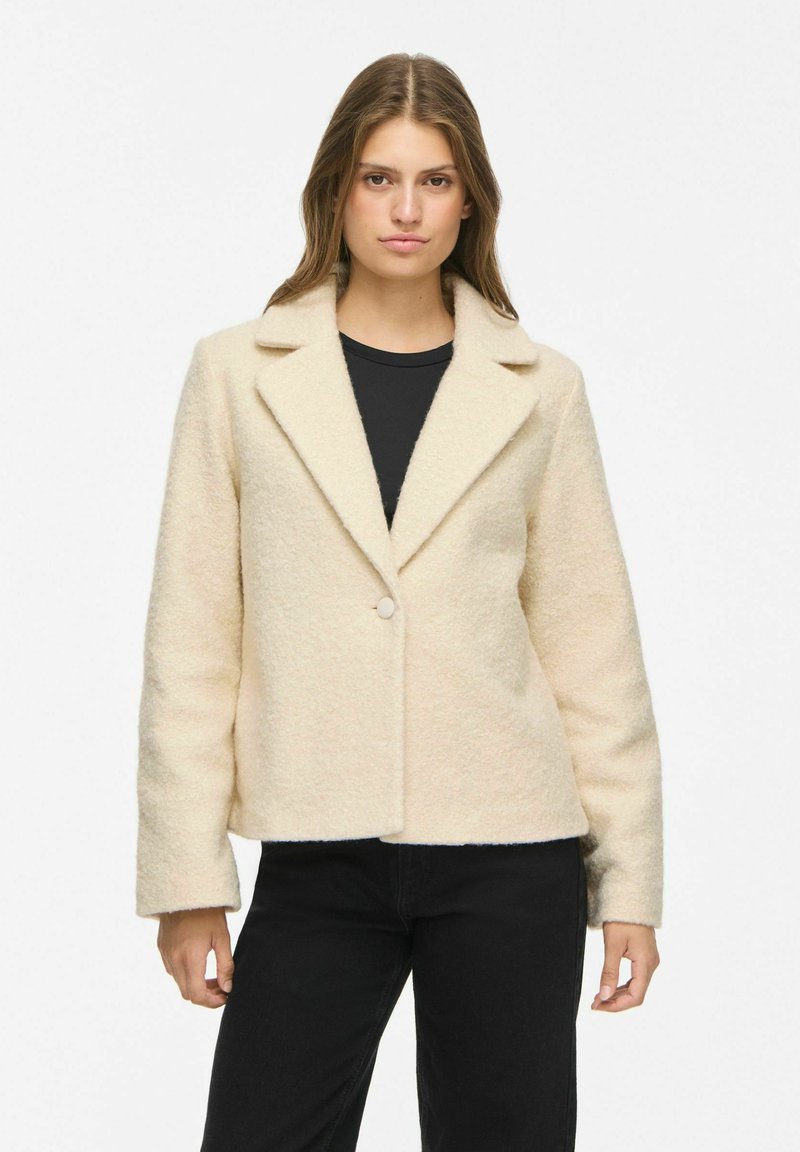 Femme portant une veste en laine texturée couleur crème avec un seul bouton, chemise noire et pantalon noir, debout devant un fond blanc uni.