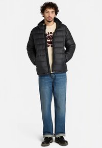 Svart vadderad jacka med dragkedja, huva och quiltad design. Bärs över en beige sweatshirt och kombineras med blå jeans och svarta skor.