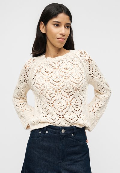 Γυναίκα με μακριά σκουρόχρωμα μαλλιά που φοράει ένα εκρού πουλόβερ με ανοιχτή πλέξη και σκούρο μπλε τζιν με ψηλή μέση, ποζάροντας με τα χέρια πίσω από την πλάτη της.