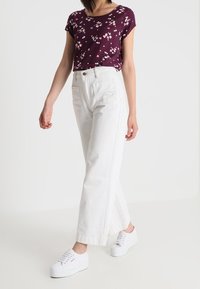 Kobieta ubrana w fioletową kwiecistą bluzkę z krótkim rękawem, białe spodnie z wysokim stanem i szerokimi nogawkami oraz białe trampki na platformie, stojąca na białym tle.