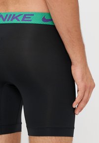 Fekete sportos rövidnadrág, textúrált zöld és lila derékkal, rajta a Nike logójával. Simább anyag, feszes illeszkedés, combközép hosszúság.