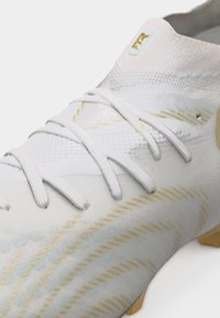 Άσπρο ποδοσφαιρικό παπούτσι με χρυσές λεπτομέρειες, υφασμένο επάνω μέρος με υφή, άσπρα κορδόνια και σόλα με χρυσά καρφιά, εμφανής σε απλό φόντο.