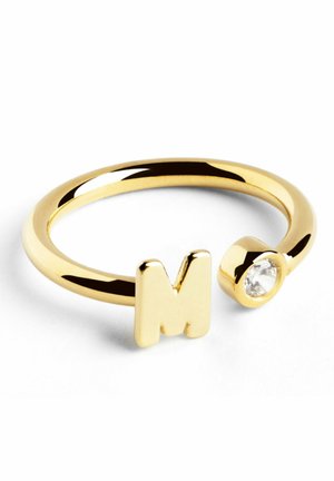 Anello in metallo dorato con lettera maiuscola "M" e un accento di cristallo trasparente. La fascia ha una texture liscia e lucida.
