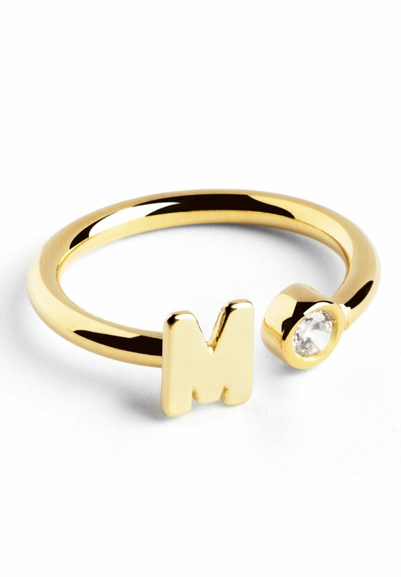Anello in metallo dorato con lettera maiuscola "M" e un accento di cristallo trasparente. La fascia ha una texture liscia e lucida.