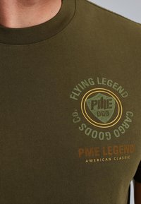 Olivno zelena bombažna majica s krožnim ovratnikom, ki vsebuje okrogel logo v temno zeleni barvi in oranžnem besedilu: "PME LEGEND AMERICAN CLASSIC."