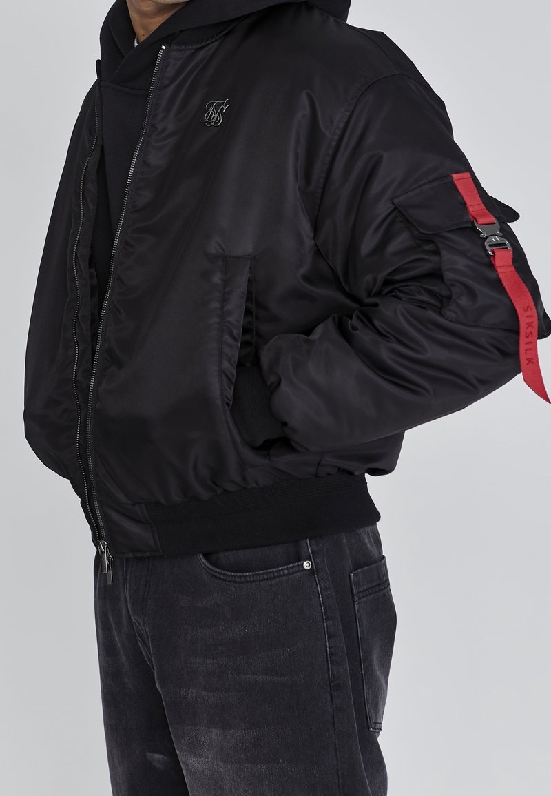 Homme portant une veste aviateur noire avec une fermeture éclair à l'avant, des poches latérales et un détail de sangle rouge sur la poche de la manche gauche, par-dessus un sweat à capuche noir et un jean.