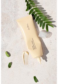 Ljus beige tub med spetsig topp och texturerat lock, märkt "Moringa All Beauty Cream," placerad på en neutral bakgrund med gröna löv.