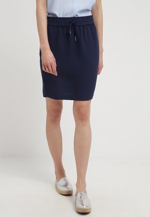 Vrouw draagt een marineblauwe rok tot op de knie met een elastische tailleband en koord, gecombineerd met zilveren instapschoenen en een lichtblauw topje.