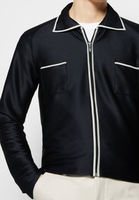  svart jacka med dragkedja och vit kant på kragen, dragkedjan och bröstfickorna, bärs över en vit skjorta med händerna i fickorna på ljusa byxor.