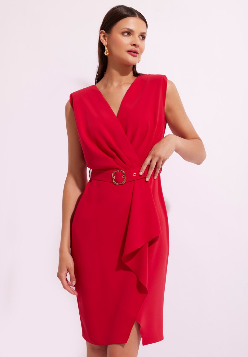 Czerwona sukienka z głębokim dekoltem w kształcie V, bez rękawów, z wcięciem w talii ozdobionym złotą klamrą oraz z plisowanym przodem. Gładka tekstura materiału.