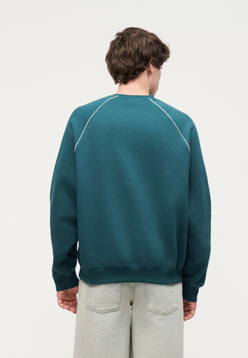 Homme portant un sweat-shirt bleu sarcelle avec un liseré blanc sur les épaules, jean gris clair, de dos contre un fond clair uni.