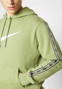 Grön hetsjacka med en stor vit Nike-logga, känguruficka och randiga detaljer med upprepande Nike-loggor på ärmarna.