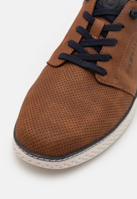 Braune, perforierte Ledersneaker mit marineblauen Schnürsenkeln, runder Zehenpartie und leichter Gummisohle, die ein geprägtes Muster entlang der Kante aufweist.