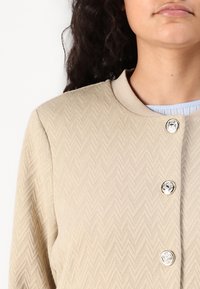 Beige strukturerad jacka med ett chevronmönster, med en rund halsringning och två glänsande knappar framtill.