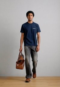 Sötétkék póló kis fehér logóval, szürke farmer, barna bőr utazótáska. A ruházathoz barna cipő tartozik.