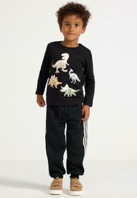 Czarny t-shirt z długim rękawem z kolorowymi nadrukami dinozaurów; połączony z ciemnymi spodniami dresowymi z białymi paskami po bokach i brązowymi butami.