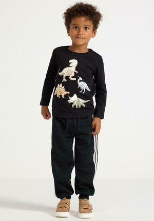Czarny t-shirt z długim rękawem z kolorowymi nadrukami dinozaurów; połączony z ciemnymi spodniami dresowymi z białymi paskami po bokach i brązowymi butami.