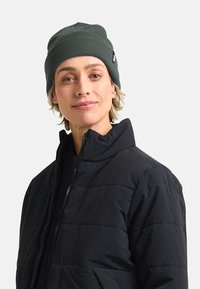 Persona con un berretto verde scuro e una giacca a vento nera con zip, che guarda in avanti con un leggero sorriso su uno sfondo chiaro e semplice.