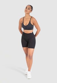 Svart sportbehå med tunna band och rynkad detalj, kombinerad med svarta cykelbyxor med hög midja. Modellen står i vita sportskor.