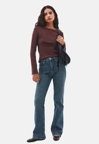 Langärmliges, kastanienbraunes Oberteil mit strukturiertem Finish, kombiniert mit hoch geschnittenen, ausgestellten blauen Jeans. Schwarze Handtasche, flache schwarze Schuhe.