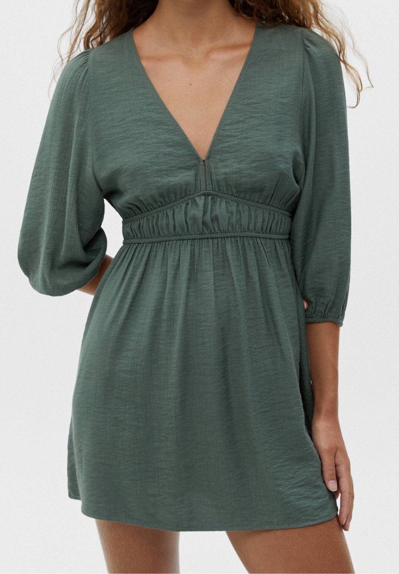 Femme portant une robe courte verte avec des manches trois-quarts bouffantes et un détail froncé à la taille, debout devant un fond blanc uni.