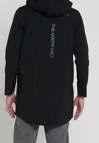Giacca nera impermeabile con cappuccio, caratterizzata da un design lungo. Sul retro è presente la scritta "THE NORTH FACE" in lettere riflettenti. Texture liscia.